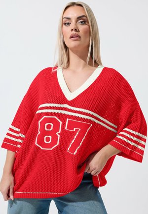 Blonde woman wearing a loose red mesh sweater with white stripes and the number 87, paired with blue jeans, against a plain background.