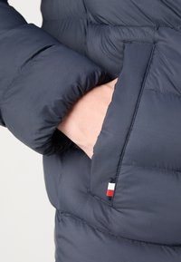 Navy insulated puffer jacket with quilted texture, featuring a side pocket and small logo detail in red, white, and blue. Soft material.