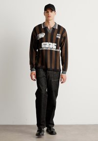 Camicia a maniche lunghe a strisce marroni e nere con colletto grigio, decorata con testo bianco. Indossata con pantaloni in denim scuri e scarpe nere.