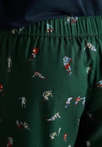 Zaļas šortas ar rakstu, kurās attēloti mazi, atkārtoti zīmējumi par rugbija spēlētājiem dažādās pozās, elastīga josta, gluda auduma tekstūra.