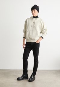 Sudadera beige con bolsillo frontal y cordones negros, combinada con jeans ajustados negros y botas negras hasta el tobillo. El modelo lleva un gorro negro.