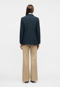 Γυναίκα που στέκεται με την πλάτη, φοράει σκούρο μπλε σακάκι, μπεζ παντελόνι με φαρδύ πόδι και μαύρα παπούτσια με τακούνι σε απλό φόντο.