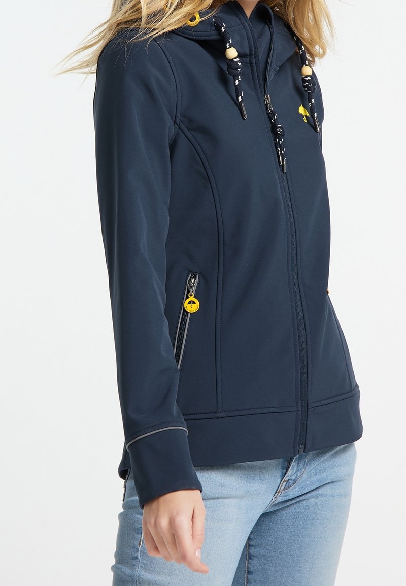 Femme portant un sweat à capuche bleu marine avec des curseurs de fermeture éclair jaunes et un jean clair, debout devant un fond blanc uni.