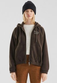 Bruine fleece zip-up hoodie met een trekkoordkap en voorzakken, gelaagd over een crèmekleurig fuzzy topje, gecombineerd met bruine broek en een zwarte beanie.