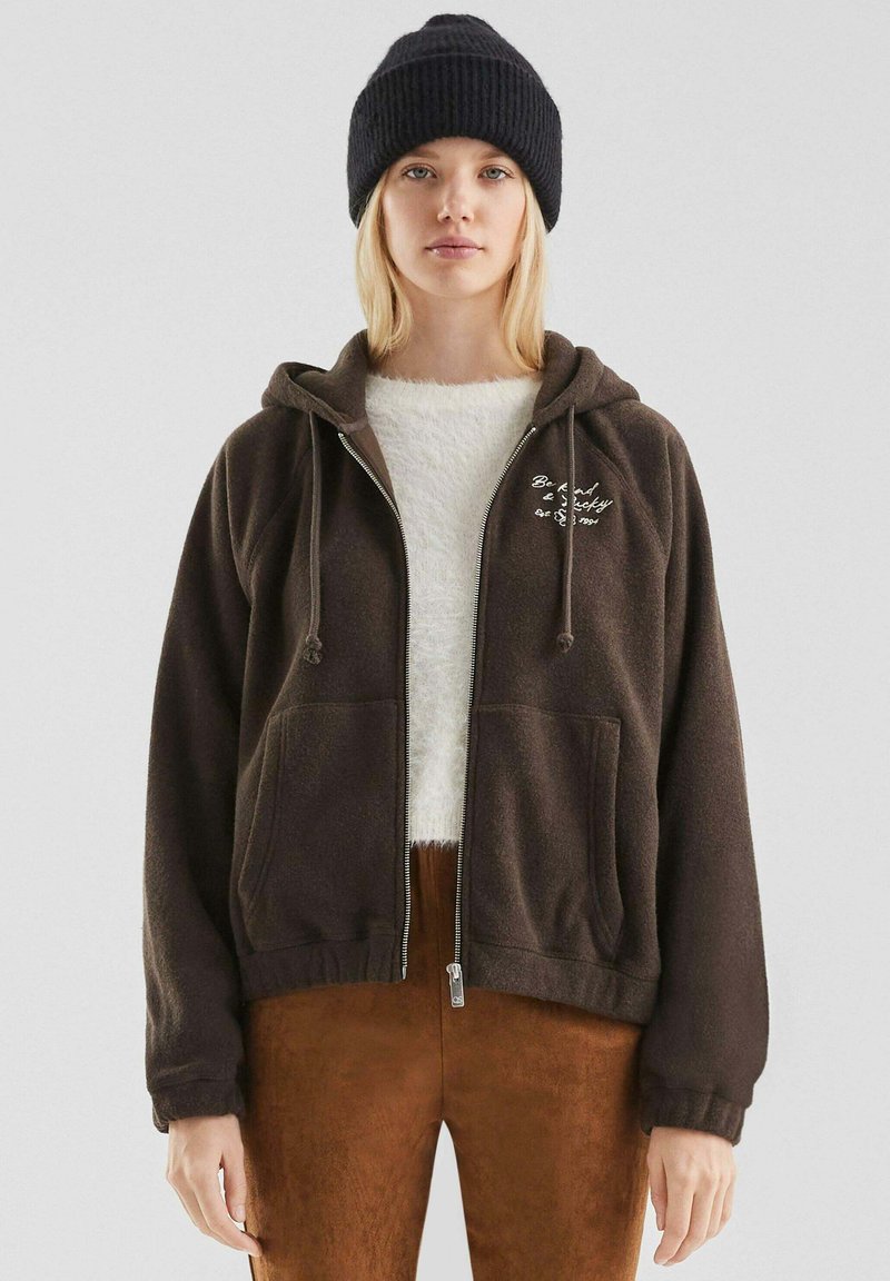 Bruine fleece zip-up hoodie met een trekkoordkap en voorzakken, gelaagd over een crèmekleurig fuzzy topje, gecombineerd met bruine broek en een zwarte beanie.