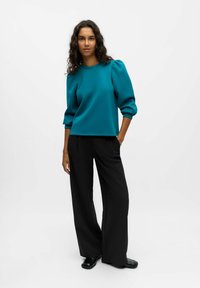 Maglia a maniche lunghe color turchese con spalle a palloncino, abbinata a pantaloni larghi neri a vita alta e scarpe nere, su uno sfondo neutro.