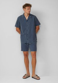 Denim overhemd met korte mouwen in donkerblauw, met knopen en een relaxte kraag; bijpassende shorts en bruine sandalen met een decoratieve strap.