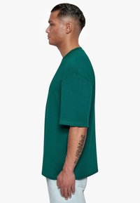 Camiseta de algodón verde con mangas cortas, un corte relajado y un escote redondo, exhibida en una persona de perfil contra un fondo blanco.