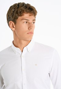 Witte button-up shirt gemaakt van katoen, met een puntige kraag en een klein gouden logo op de linkerkant van de borst. Gladde textuur en klassieke pasvorm.