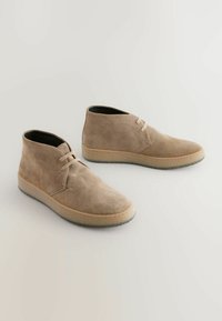 Bottines en daim beige avec bout rond, lacets et semelle en caoutchouc texturée. Finition lisse avec des coutures légères contrastantes autour du bord.