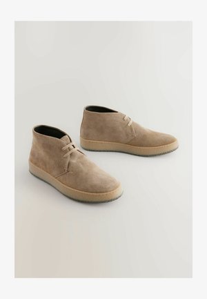 Beige Wildleder-Stiefeletten mit abgerundeter Spitze, Schnürung und strukturierter Gummisohle. Glatte Oberfläche mit kontrastierenden hellen Nähten entlang der Kante.
