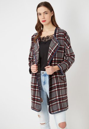 Abrigo de cuadros con base gris y acentos en rojo y azul, diseño de frente abierto, grandes bolsillos y textura suave. Lleva puesto un top negro.