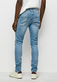 Man wearing light blue slim-fit jeans and white sneakers with maroon heel detail, standing against a plain white background.