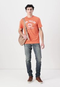 Camiseta de algodón naranja con estampado gráfico en blanco, combinada con jeans azul deslavado y zapatillas marrones. Se lleva un bolso de hombro beige estilo bandolera.