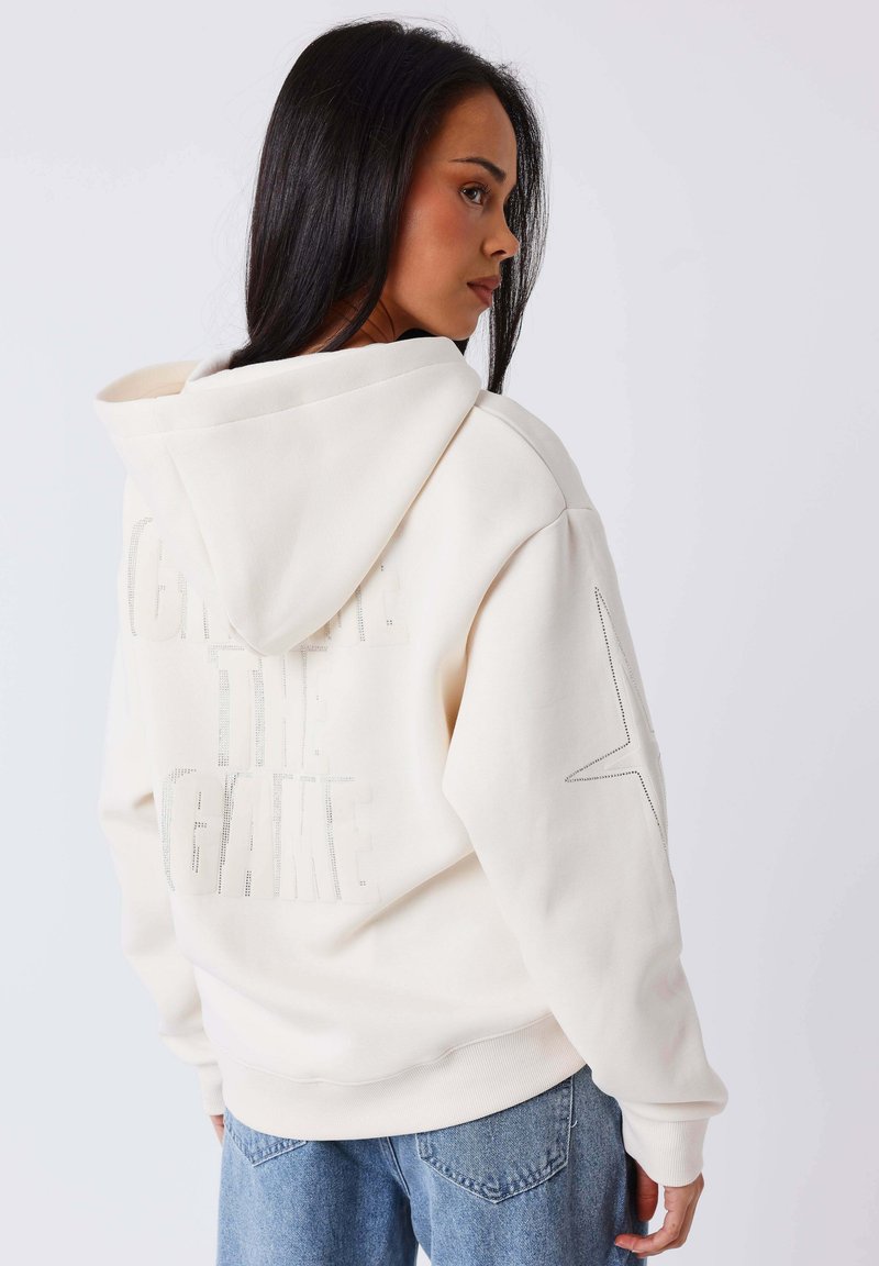 Femme aux longs cheveux noirs portant un sweat à capuche blanc avec un texte perforé et un motif d'étoile dans le dos, vêtue d'un jean bleu, vue de profil.