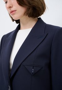 Marinblå blazer med skarpa slag, enkel knäppning och en triangulär fickdetalj. Tillverkad av strukturerad tyg med en slät yta.