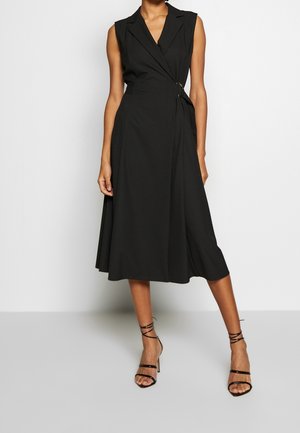 Sleeveless black midi wrap dress with lapel collar, secured with a side metal ring, worn with black strappy high heels.
