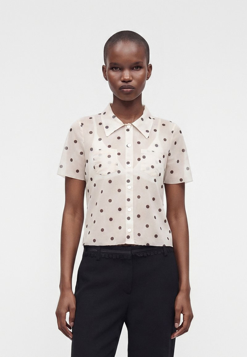 Femme portant une chemise beige transparente à manches courtes boutonnée avec des pois noirs et un pantalon noir, debout devant un fond blanc uni.
