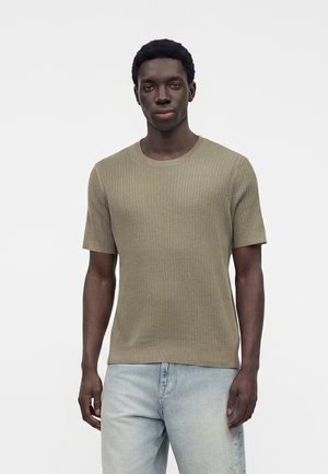 Junger Mann trägt ein hellbraunes, geripptes Kurzarmshirt und hellblaue Jeans und steht vor einem schlichten weißen Hintergrund.
