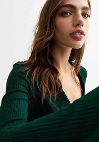 Abito verde a coste con maniche lunghe e scollatura a V profonda, che mette in risalto un tessuto texturizzato. I capelli appaiono mossi e castani, incorniciando il viso.