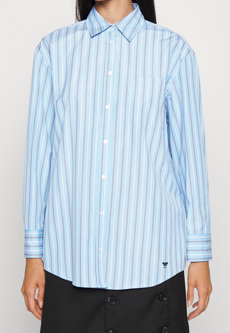 Personne portant une chemise ample à manches longues, rayée verticalement bleu et blanc, avec une poche poitrine et une jupe noire.