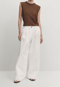 Bruine geribbelde mouwloze top gedragen met hoge witte wijde broek met knoopdetails en plooien. Zwarte sandalen.