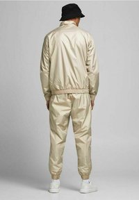 Beige nylon träningsoverall med elastiska mudd och midjeband, med en blank finish. Bärs med en svart bucket hat och vita sneakers.