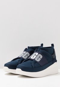 Granatowe buty sportowe z elastyczną cholewką z materiału, elastycznym paskiem z wyraźnym szarym logo oraz białą, grubą gumową podeszwą.