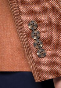 Brązowy materiał kurtki w wzory z teksturowanym splotem, z czterema okrągłymi, ciemnymi guzikami na mankiecie.