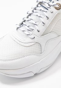 Baskets blanches avec une tige en mesh texturé, des accents en cuir lisse et des lacets à motifs contrastants. Semelle incurvée avec coutures renforcées.