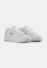 Adidași albi cu partea superioară din piele netedă, vârf rotund și talpă din cauciuc plat. Au detalii cusute și un logo negru pe lateral.