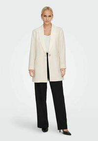 Blazer long beige avec revers crantés, fermeture à un bouton et deux poches avant, associé à un pantalon noir à jambes larges et des talons.