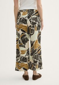 Kobieta nosząca luźne, szerokie spodnie z dużym czarnym, brązowym i beżowym wzorem liści, w zestawieniu z brązowymi płaskimi butami, pokazana z tyłu.