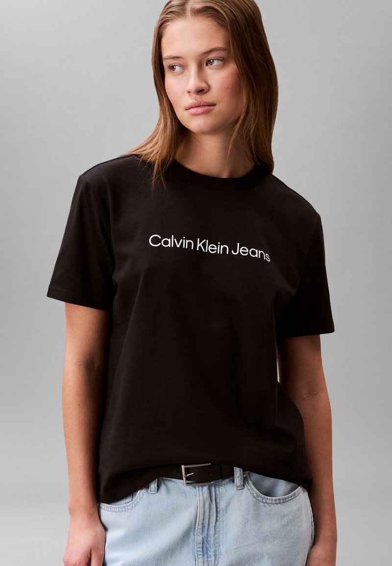 Czarny bawełniany t-shirt z białym logo "Calvin Klein Jeans", krótkie rękawy, okrągły dekolt, zestawiony z jasnoniebieskimi dżinsami.