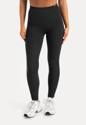 Vrouw die zwartelegging met hoge taille en witte sportschoenen draagt, staand tegen een effen lichte achtergrond.