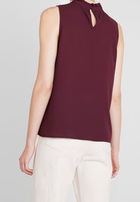 Top senza maniche color bordeaux con colletto alto e dettagli arricciati, caratterizzato da un'apertura a chiave sul retro. Tessuto morbido, vestibilità rilassata, abbinato a pantaloni di colore chiaro.