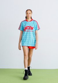 T-shirt azul claro de mangas curtas com detalhes vermelhos e um estampado gráfico, combinado com calções laranja e botas pretas de atacador num chão verde.