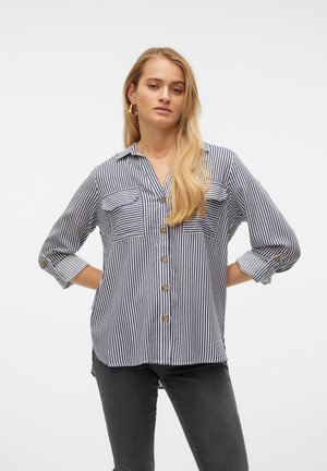 Femme aux longs cheveux blonds portant une chemise boutonnée à rayures verticales bleu marine et blanc et un jean foncé, debout, les mains sur les hanches.