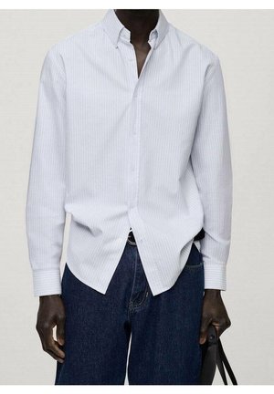 Mężczyzna ubrany w białą koszulę w prążki, wpuszczoną w ciemnoniebieskie dżinsy, trzymający czarną torbę w prawej ręce na jednolitym tle.