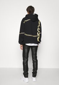 Felpa nera con stampa di catene dorate e logo sul retro, abbinata a pantaloni in pelle nera aderenti e sneakers bianche.