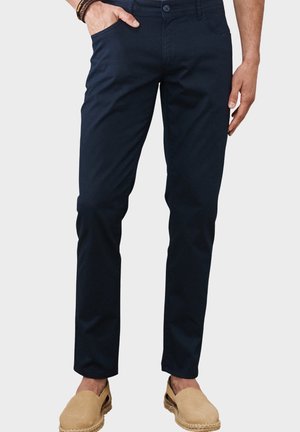 Mand iført mørkeblå tætsiddende bukser med beige slip-on sko, stående med den ene hånd i lommen mod en ensfarvet baggrund.