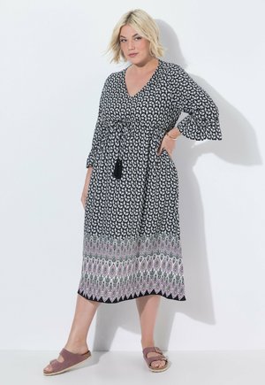 Kvinde med kort blond hår iført en sort-hvid mønstret midikjole med mønstret kant og lyserøde slip-on sandaler, poserer med den ene hånd på hoften.