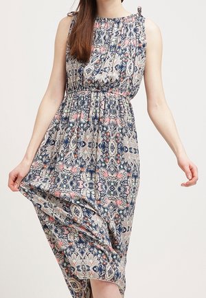 Kvinna som bär en ärmlös, ankellång klänning med blå, rosa och beige blommönster och geometriska mönster, håller fållen lätt lyft.