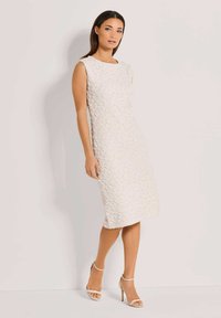 Ärmelloses knielanges Kleid aus strukturiertem cremefarbenem Stoff mit einem Blumenmuster. Kombiniert mit hellen hohen Sandalen.