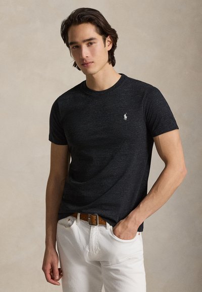 Sort crewneck T-shirt i tekstureret stof med en lille hvid logo på venstre bryst, kombineret med hvide jeans og et brunt bælte.