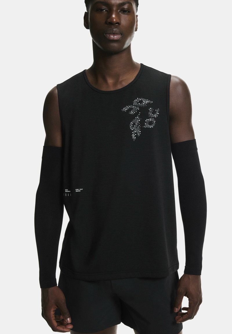 Jeune homme portant un haut noir sans manches avec un motif de feuille argentée et des manches noires, debout devant un fond clair uni.