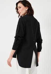 Mustan nappipaidan malli, jossa on korkea matala design, pitkät hihat ja hihansuut sekä pyöristetty helma. Pehmeää kangasta, jolla on sileä pinta.