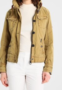 Beige bomulljacka med huva, flera framfickor, knapplikning och dragkedja, samt justerbara ärmslut, bärs över en ljus ribbad topp.