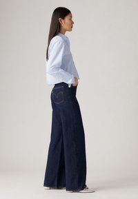 Femeie stând de profil, cu părul lung și închis la culoare, purtând o cămașă butonată de culoare albastră deschisă, blugi largi de culoare închisă și pantofi albi cu vârful ascuțit.