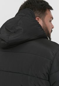 Sort pufferjakke med hætte. Har syede paneler og en glat, let skinnende tekstur. Bagfra ses en afrundet krave.